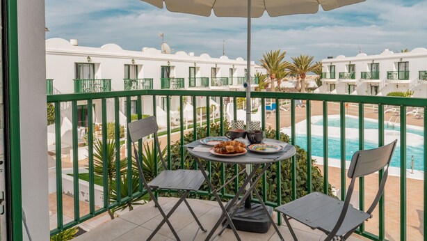 Un balcon avec une table, des chaises et un parasol, donnant sur une piscine et les bâtiments de l’hôtel.