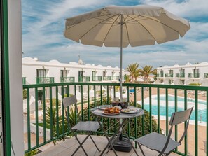 A balcony with a table, chairs, and an umbrella overlooking a pool and hotel buildings. - Wohnung "El Encanto" mit Terrasse by Interhome (Corralejo)