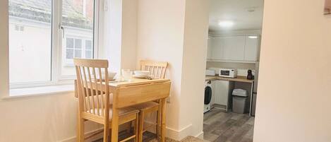 A compact living space with a dining area featuring a wooden table and chairs, a small kitchen with basic appliances, and a view through an open doorway.