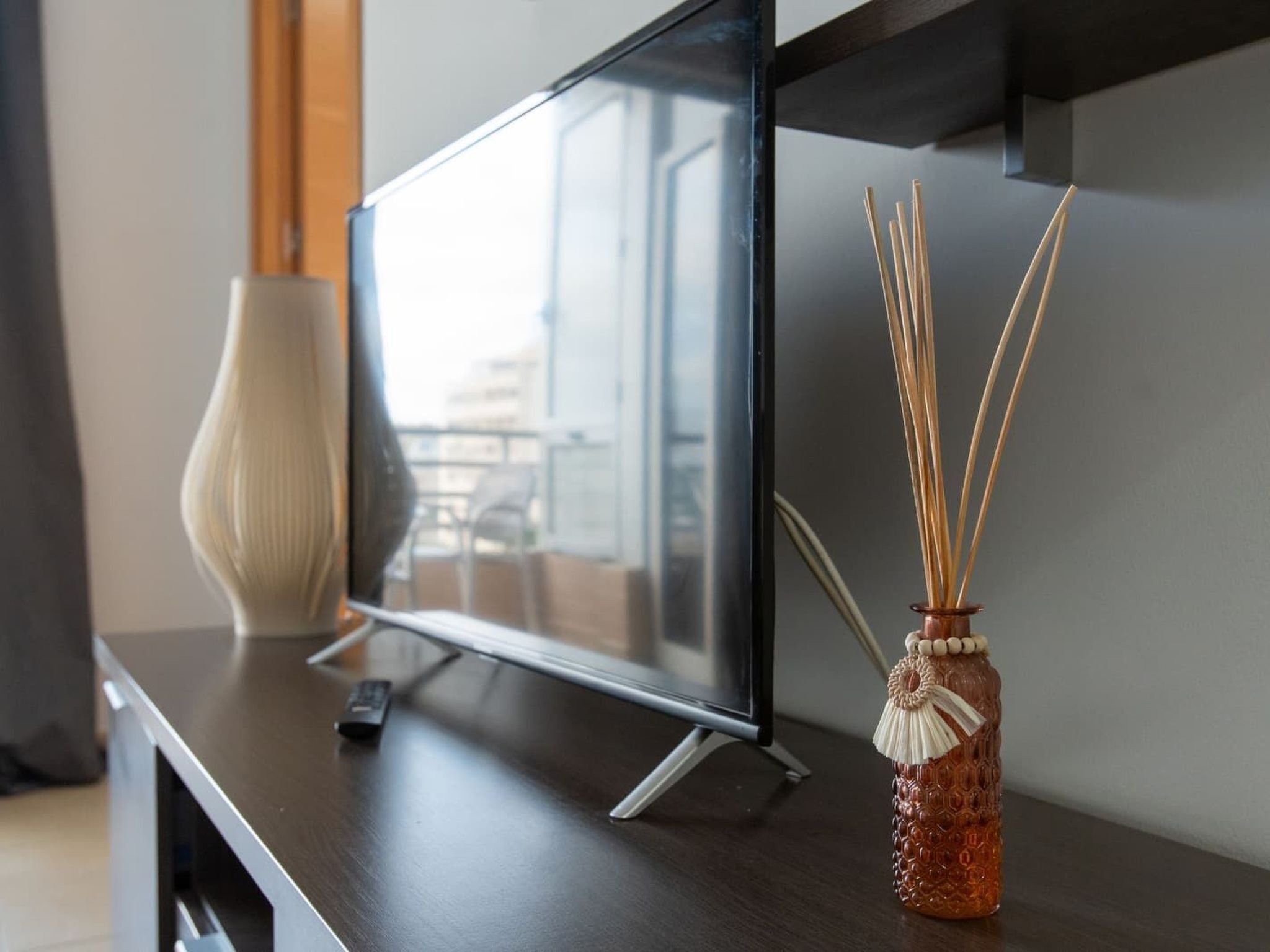 A flat-screen TV on a wooden stand, a vase with reed sticks, and a remote control on a dark surface.