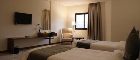 A hotel room with two beds, a desk, a chair, a TV, and a window with curtains.