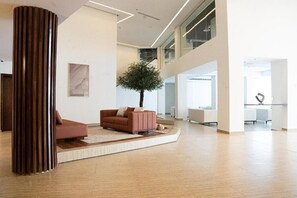 Un vestíbulo amplio con diseño moderno, que presenta una gran columna de madera, una zona de estar con sofás, un árbol central y vistas al piso superior.