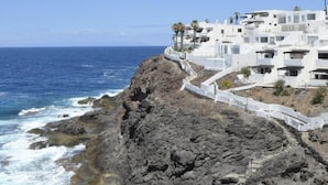 A coastal building with white walls and balconies, situated on a rocky cliff overlooking the sea.