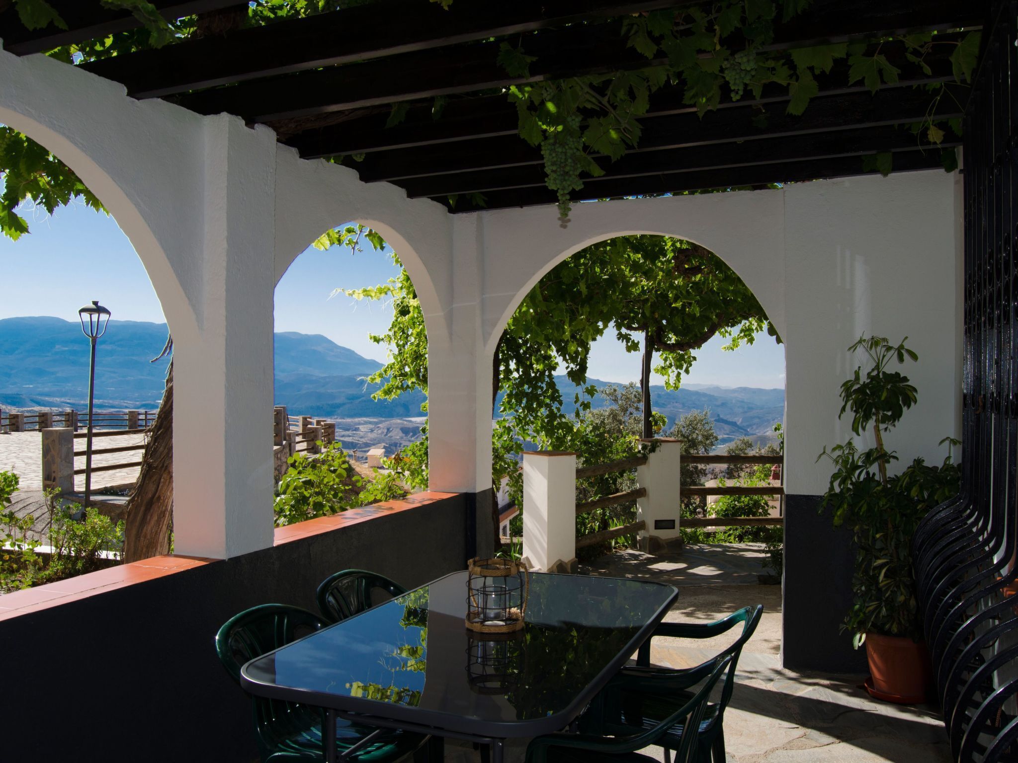 An outdoor patio with a glass table and chairs, surrounded by white arches and greenery, overlooking a scenic landscape.