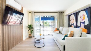A modern living room with a flat-screen TV, a sofa with cushions, a small round table with a vase, and a large window with a view of the outside. - Home2Book Stylish Studio by the Sea Los Cristianos (Los Cristianos)