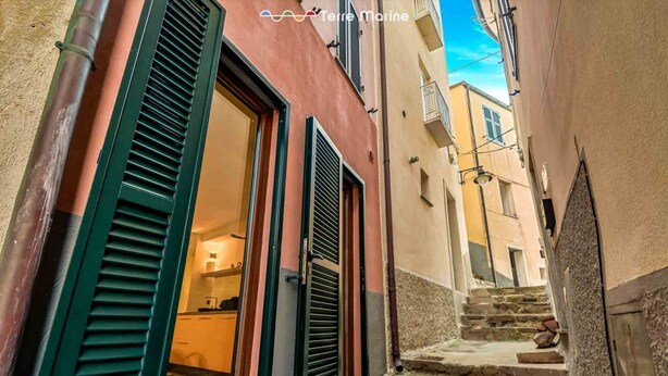 Un stretto vicolo con una scala in pietra che conduce a un edificio con persiane verdi e un interno visibile.