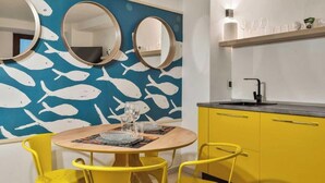 Zona pranzo con un tavolo rotondo, sedie gialle e una parete blu decorata con motivi a pesce. Si notano tre specchi rotondi sulla parete, un rubinetto nero e uno scaffale con bicchieri.