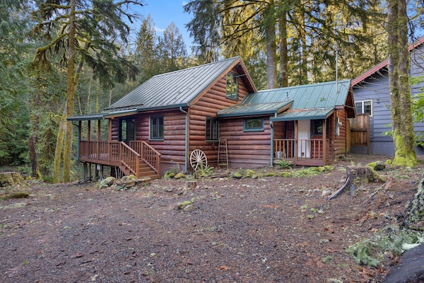 Charming log cabin nestled among towering evergreens with rustic wagon wheel detail and spacious deck for forest retreat relaxation.