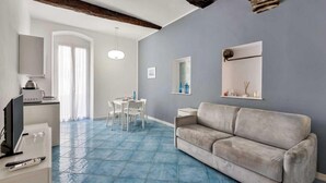 Habitació amb un sofà gris, taula de dinar amb cadires, televisió i zona de cuina amb microones.