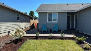 Ein Wohngebäude mit überdachter Terrasse, zwei blauen Adirondack-Stühlen und einem kleinen Gartenbereich mit weißen Blüten.