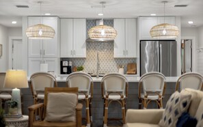 A modern kitchen with white cabinets, a stainless steel refrigerator, wicker chairs, and pendant lights.