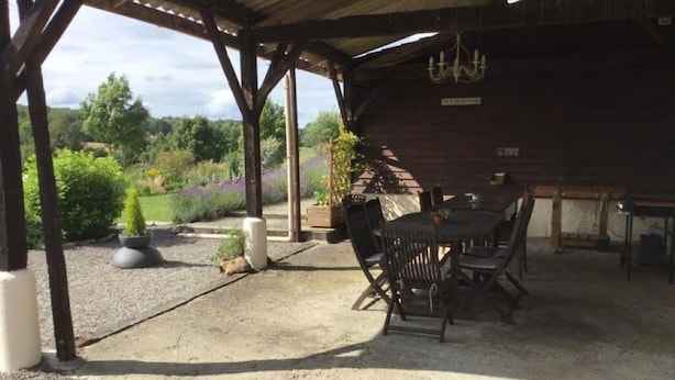A covered outdoor dining area with a rustic wooden pergola, a table with chairs, and a view of the surrounding garden and trees.