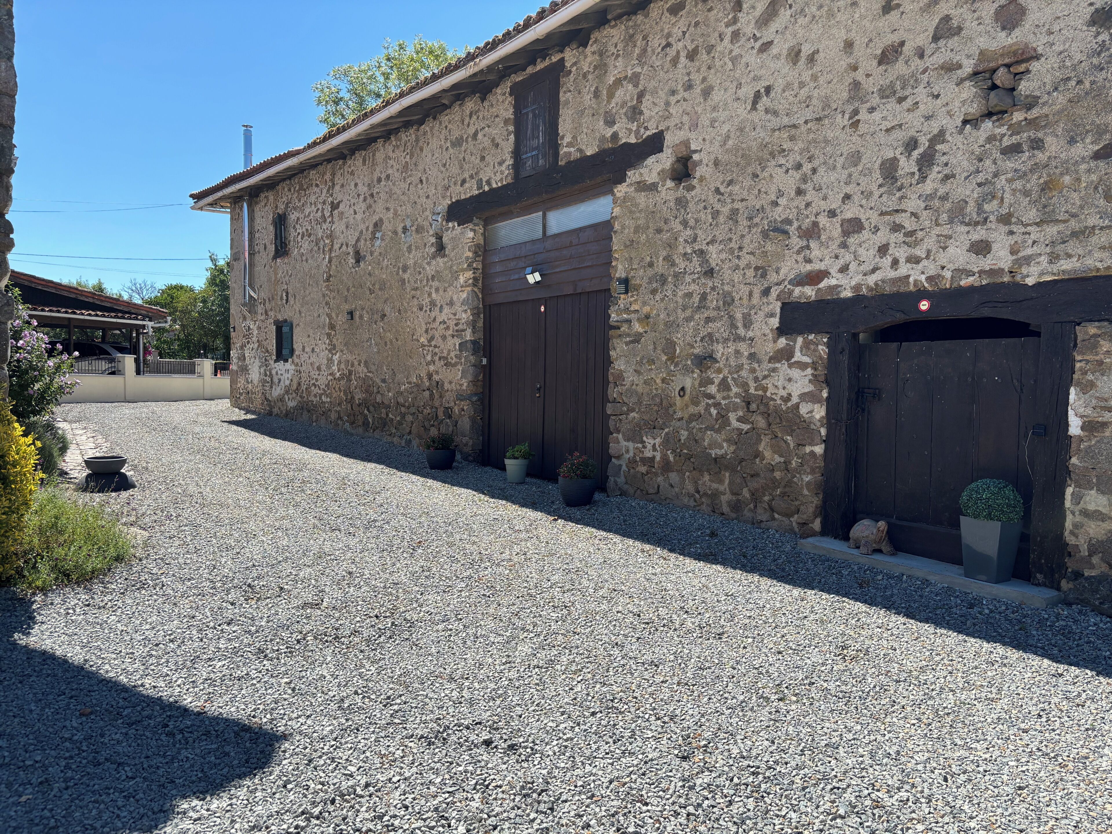 A stone building with two dark wooden doors, a gravel driveway, and a small garden area with potted plants.