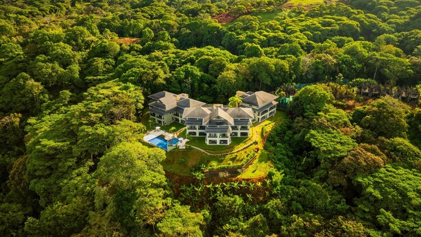Vista aérea de una amplia casa moderna rodeada de un bosque denso, con una piscina y varios edificios.