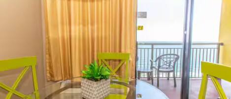 A room with a glass table, two yellow chairs, a potted plant, and a sliding glass door leading to a balcony with chairs.