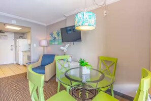 A compact living space with a dining area featuring a glass table and green chairs, a sofa, a TV mounted on the wall, and a small kitchen area. - Charming condo in quaint Myrtle Beach perfect for your getaway (Myrtle Beach)