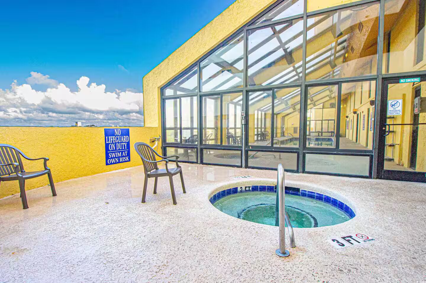 An outdoor swimming pool with a no lifeguard on duty sign, two chairs, and a clear view of the pool area through large windows.