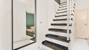 Una escalera moderna con diseño minimalista, un espejo grande que refleja una habitación con cama y pared verde, y una puerta blanca al final.
