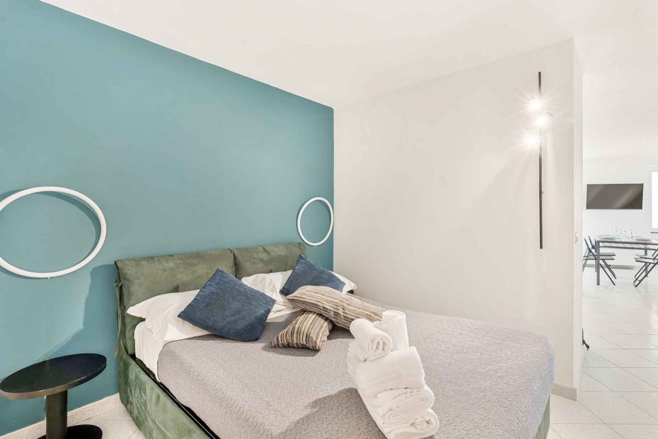 Et moderne hotelværelse med en teal farvet accentvæg, en seng med et gråt tæppe, flere puder, et lille rundt bord og en høj gulvlampe.