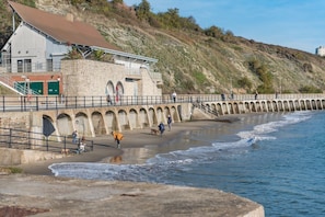 A coastal building with a stone facade, a staircase leading to a balcony, and a beach area with a low wall and a pier extending into the water. - Folkestone Apartments, Kent (Folkestone)
