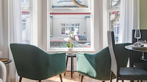 A room with two green chairs, a small table, and a view of a street through large windows.