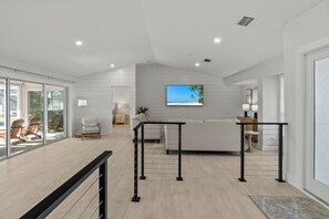 Uma sala de estar moderna com um sofá branco, uma TV na parede e uma porta de vidro que dá para uma área externa com cadeiras.
