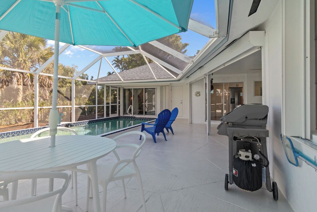 Un espacio al aire libre techado con piscina, mesa y sillas blancas, sillas azules y un carrito para asar.