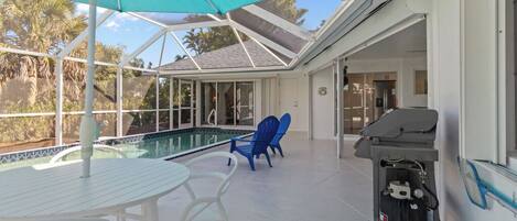 Un espacio al aire libre techado con piscina, mesa y sillas blancas, sillas azules y un carrito para asar.