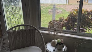 A wicker chair with a white cushion, a small round table with a plant, and a view of a lake with buildings and greenery. - Waterfront Views from Inside Home, Screened Porch & Private Deck (Tavares)