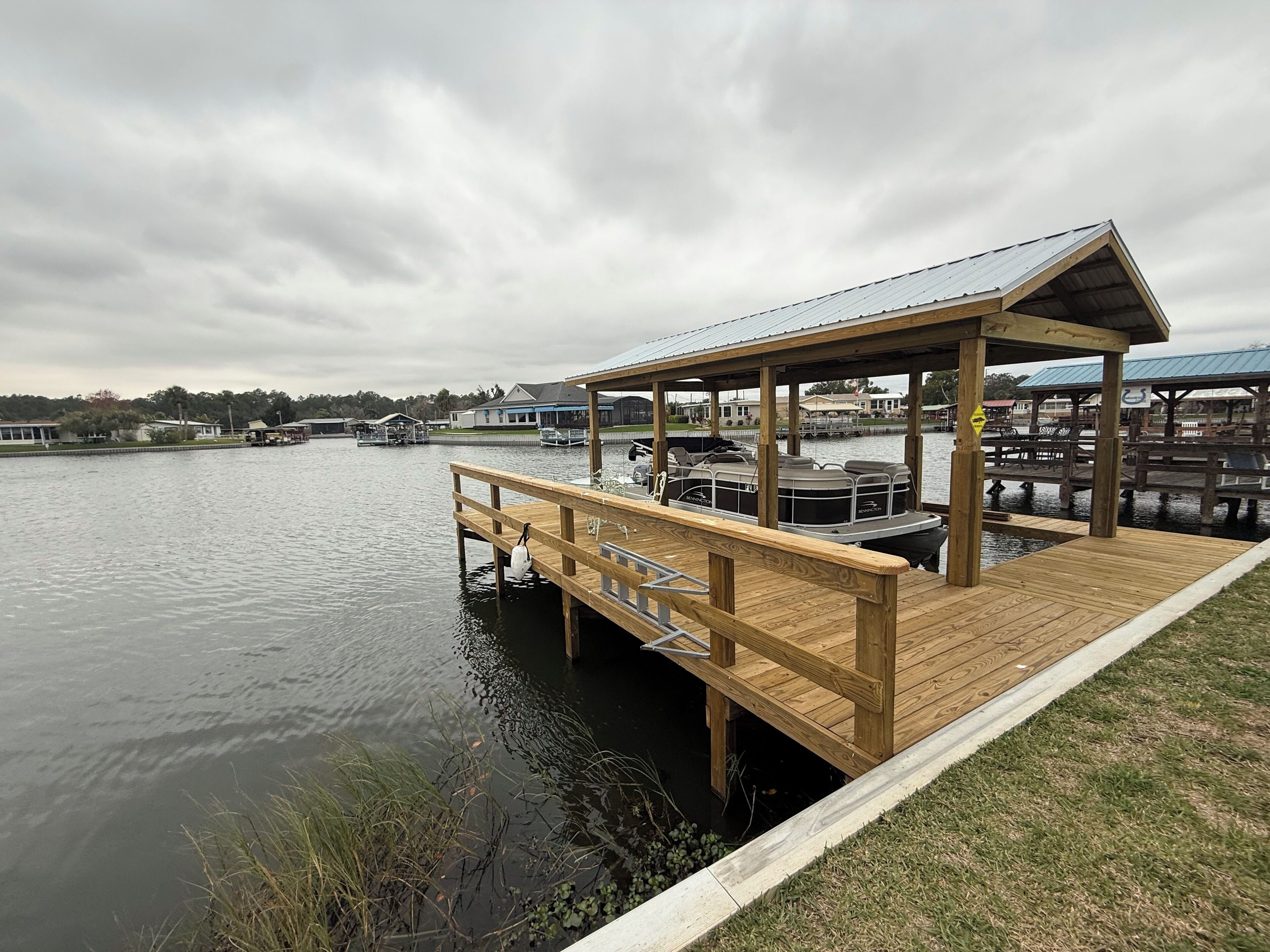 Un muelle de madera que se adentra en un lago tranquilo, con una zona de estar cubierta y un barco amarrado cerca.