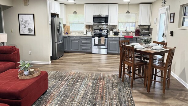 A modern kitchen with white cabinets, stainless steel appliances, and a dining area with wooden chairs and a table set for four. - Waterfront Views from Inside Home, Screened Porch & Private Deck (Tavares)