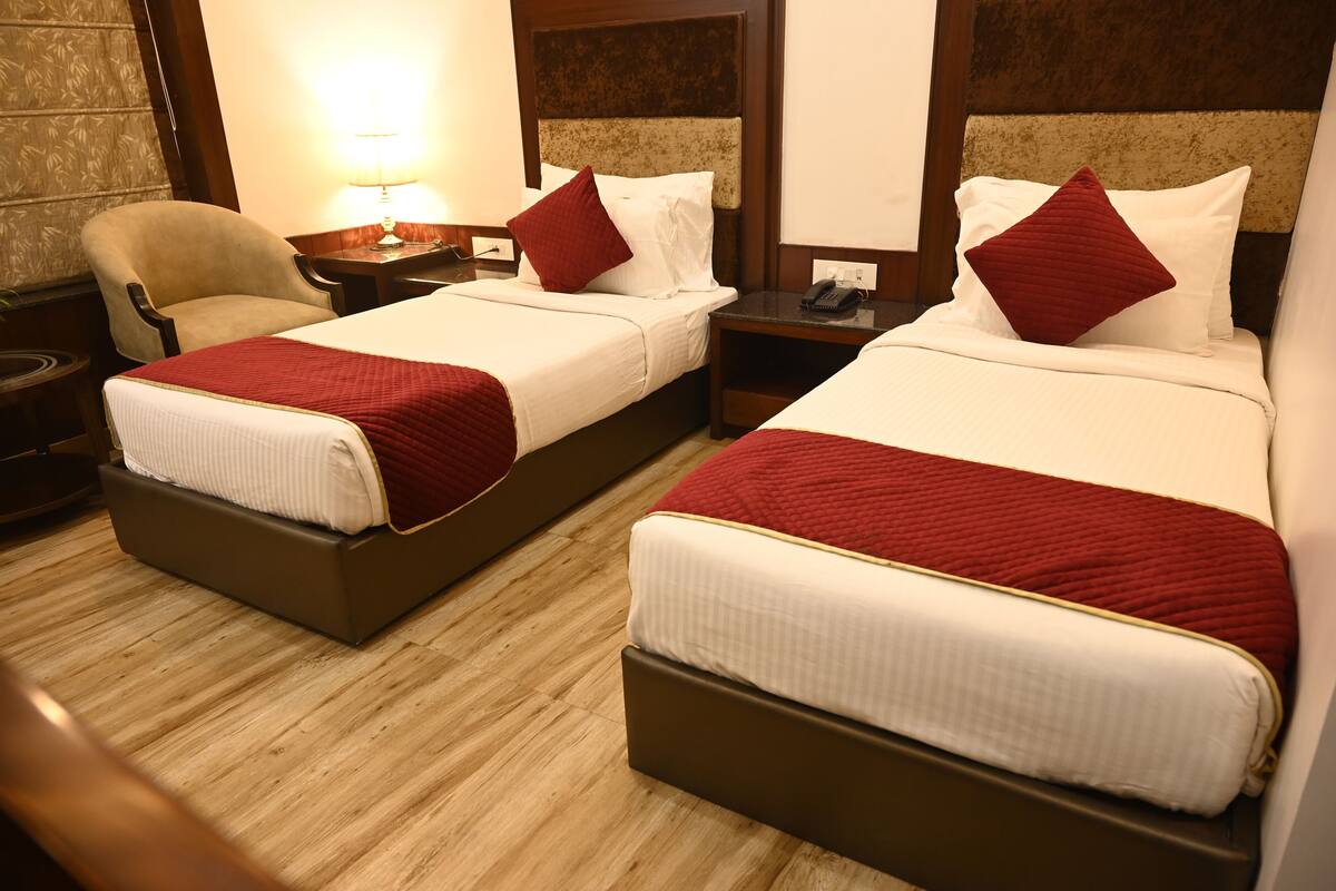 A hotel room with two beds, each with a red blanket and pillows, a wooden floor, a chair, a nightstand with a lamp, and a wall with a floral pattern.