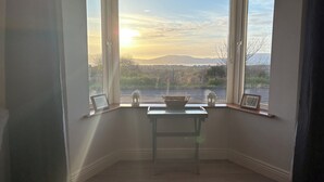 Een kamer met erker, met uitzicht op een landschap, een tafeltje met een schaal en lantaarns, en foto's in lijsten op de vensterbank.