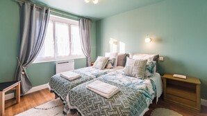 Ein Schlafzimmer mit zwei Betten, einem Fenster mit Vorhängen, Nachttischlampen, einem Nachttisch und einem Heizkörper.
