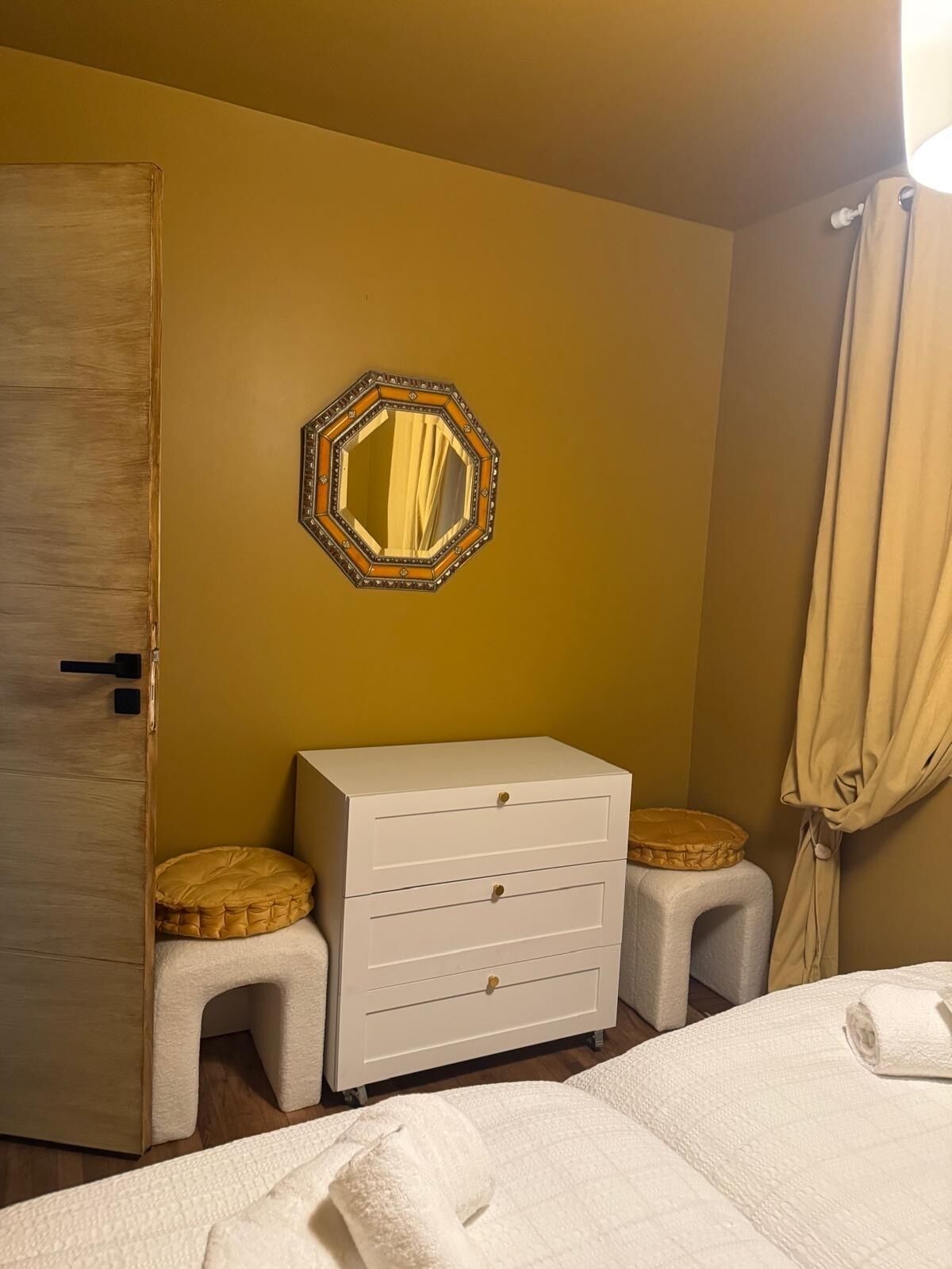 A room with a wooden door, a white dresser with drawers, two small stools, a mirror with a decorative frame, and a bed with a rolled-up towel.