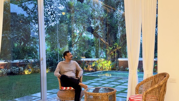 A man sitting on a wicker chair in a room with a large glass door leading to a garden. - A 3BHK Luxury Villa with Pvt Pool, Garden Near Beach (Siolim Goa)