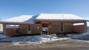 A building with a snow-covered roof and stone walls, featuring a wooden door and a bench outside. - Le Balcon du Lac - Navette devant résidence (Les Angles)