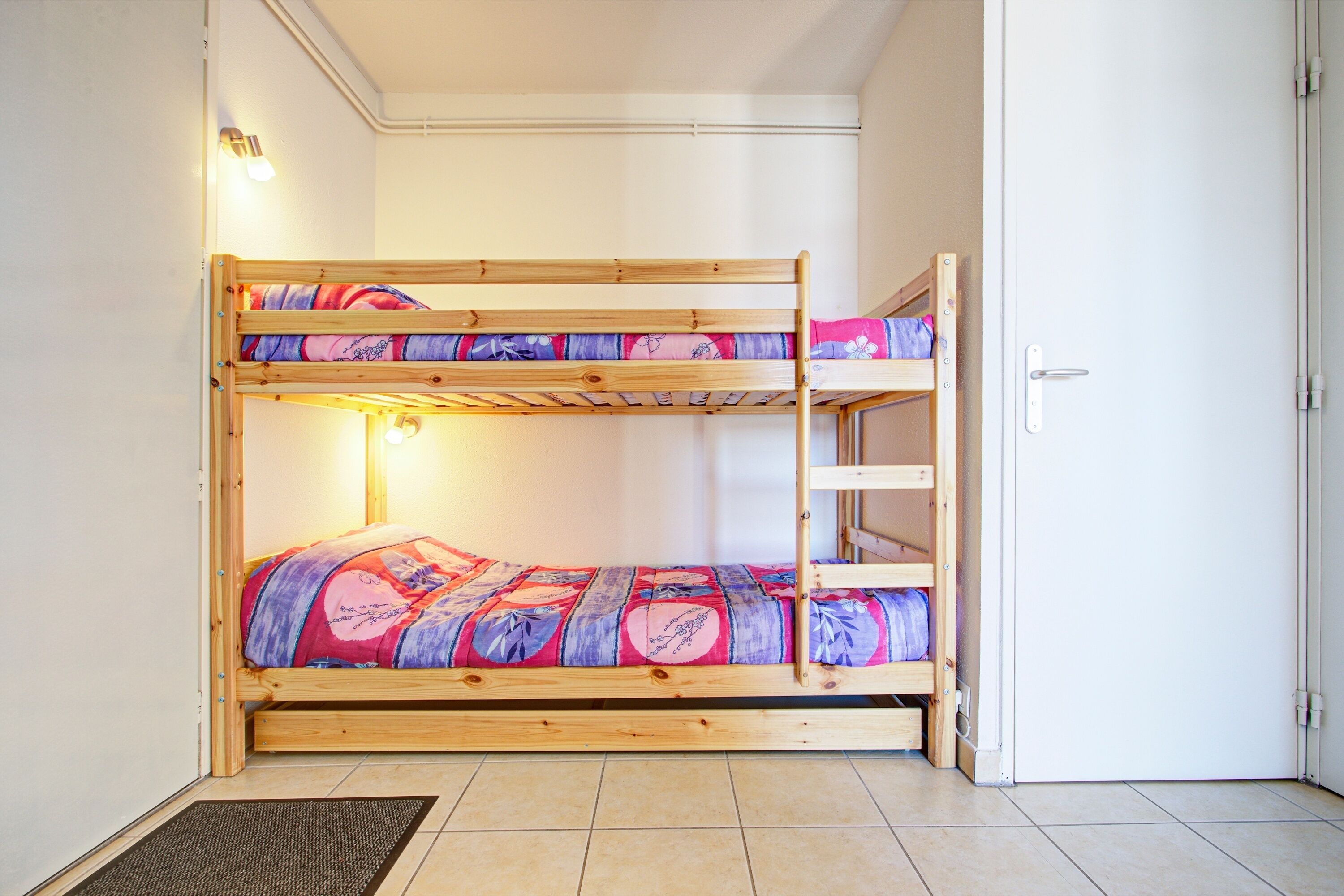 Una cama litera con un edredón de colores, una escalera y una puerta a la derecha.