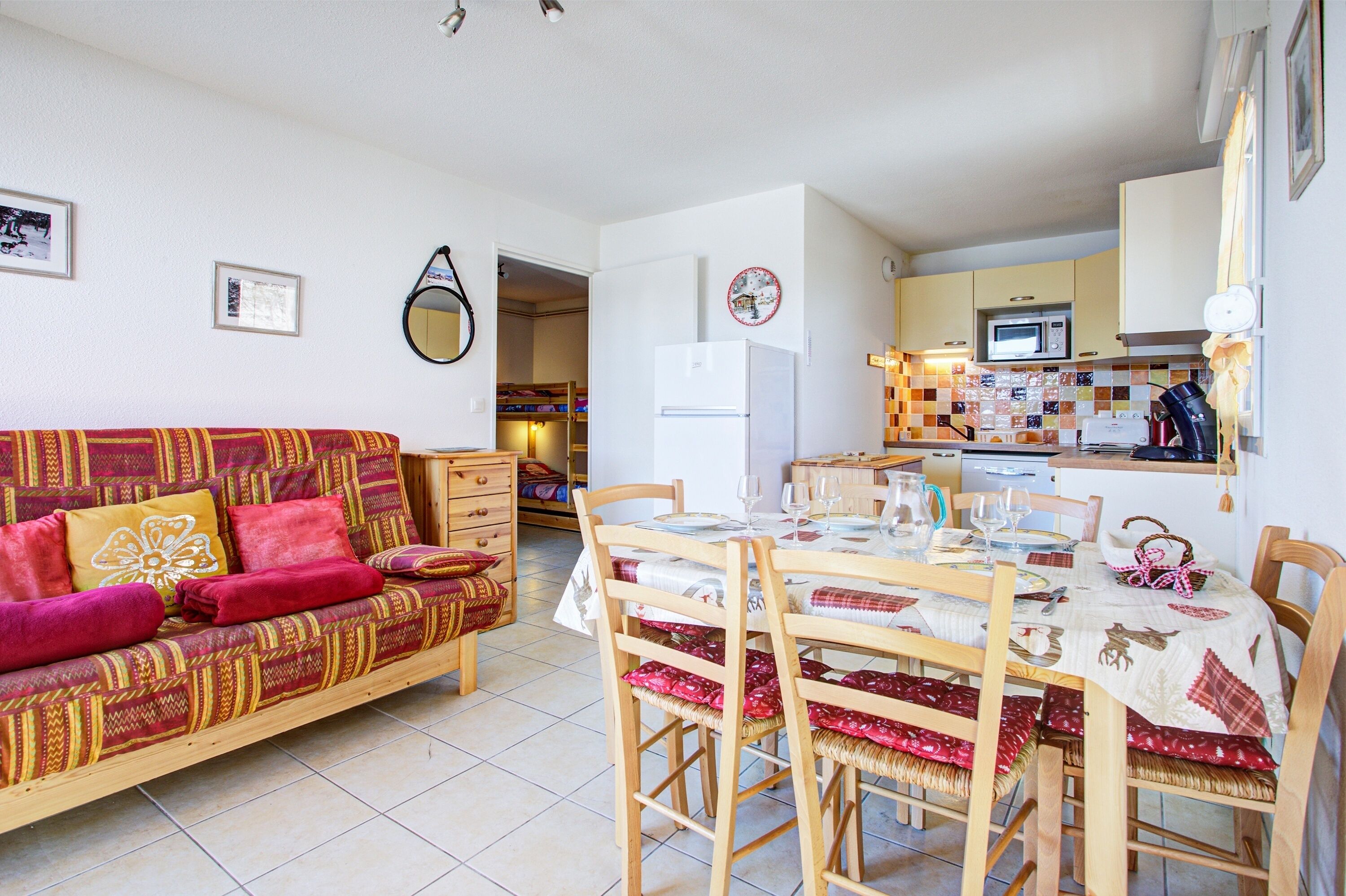 A cozy living room with a sofa, dining table, and chairs. A kitchen with a microwave and refrigerator is visible in the background.