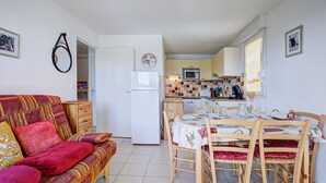 A cozy living room with a sofa and a dining table set for four. A kitchen with a refrigerator, microwave, and cabinets is visible in the background. - Le Balcon du Lac - Navette devant résidence (Les Angles)