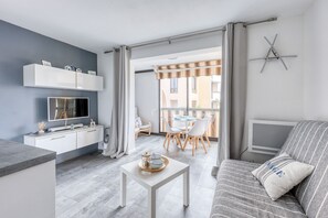 Ein modernes Wohnzimmer mit einer Couch, einem Couchtisch und einem Fernseher. Es gibt einen Essbereich mit Stühlen und einem Tisch. Der Raum verfügt über einen Balkon mit Vorhängen.