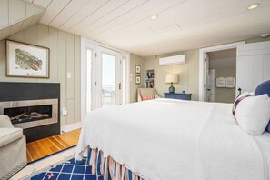 A bedroom with a large bed, a fireplace, a door leading to a balcony, and a small side table with a lamp. - Luxury 4 Bed, 3.5 Bath Oceanfront Home, KBIA, Mother’s Beach (Kennebunk)
