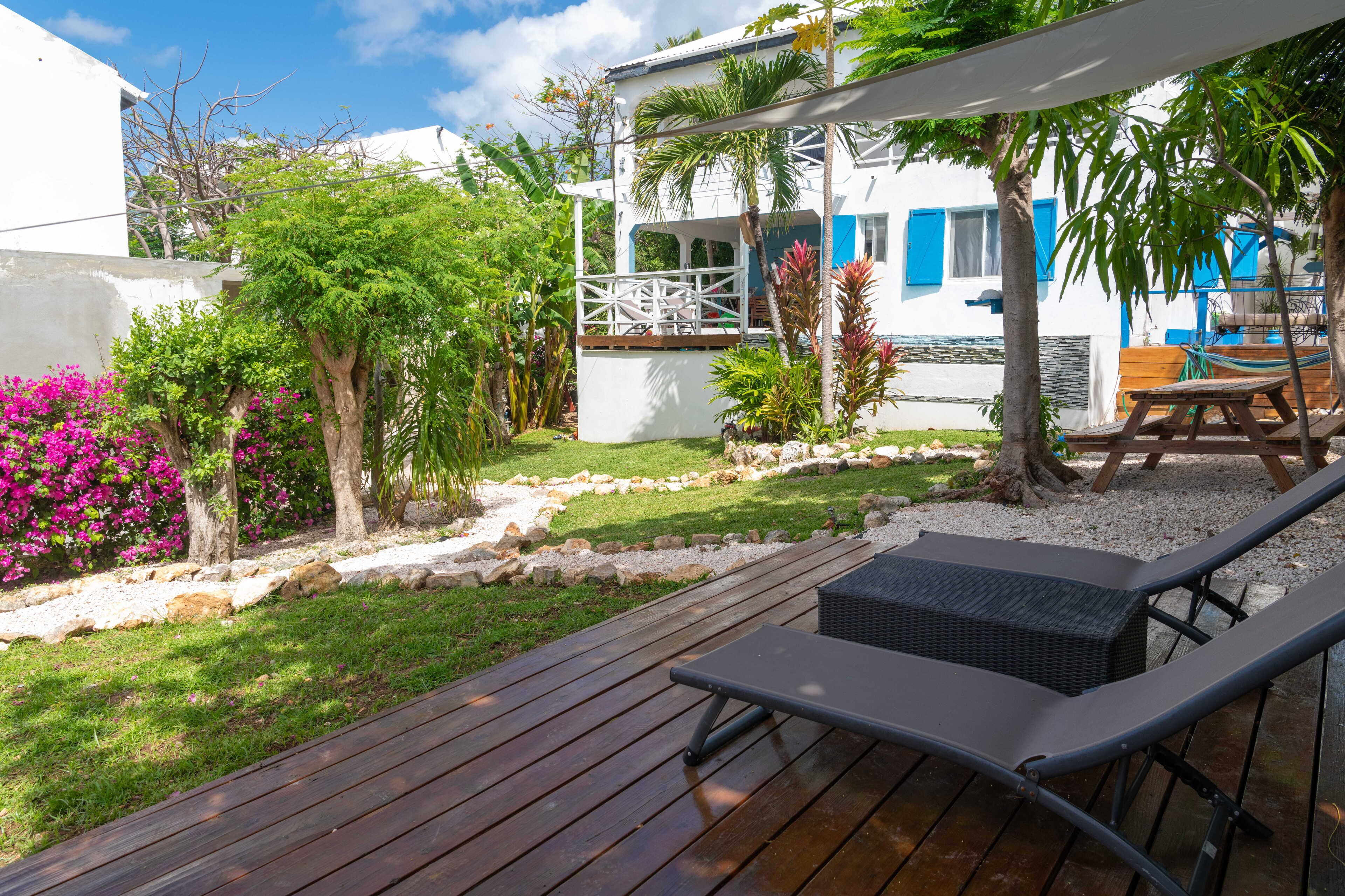 A wooden deck with lounge chairs, a white building with blue shutters, and a garden with pink flowers and trees.