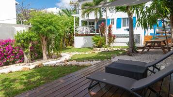 A wooden deck with lounge chairs, a white building with blue shutters, and a garden with pink flowers and trees.
