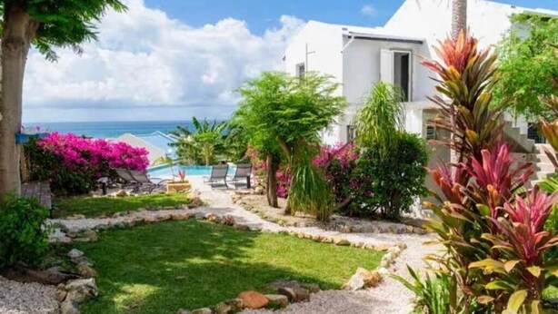 Un edificio bianco con piscina, circondato da un giardino fiorito di fiori rosa e vista sul mare.