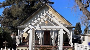 A small, single-story house with a white picket fence, a brick pathway leading to the front door, and a porch with white curtains.