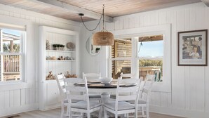 Un espace repas avec une table ronde, des chaises blanches, un lustre suspendu en osier et une vue sur l’extérieur grâce à de grandes fenêtres.