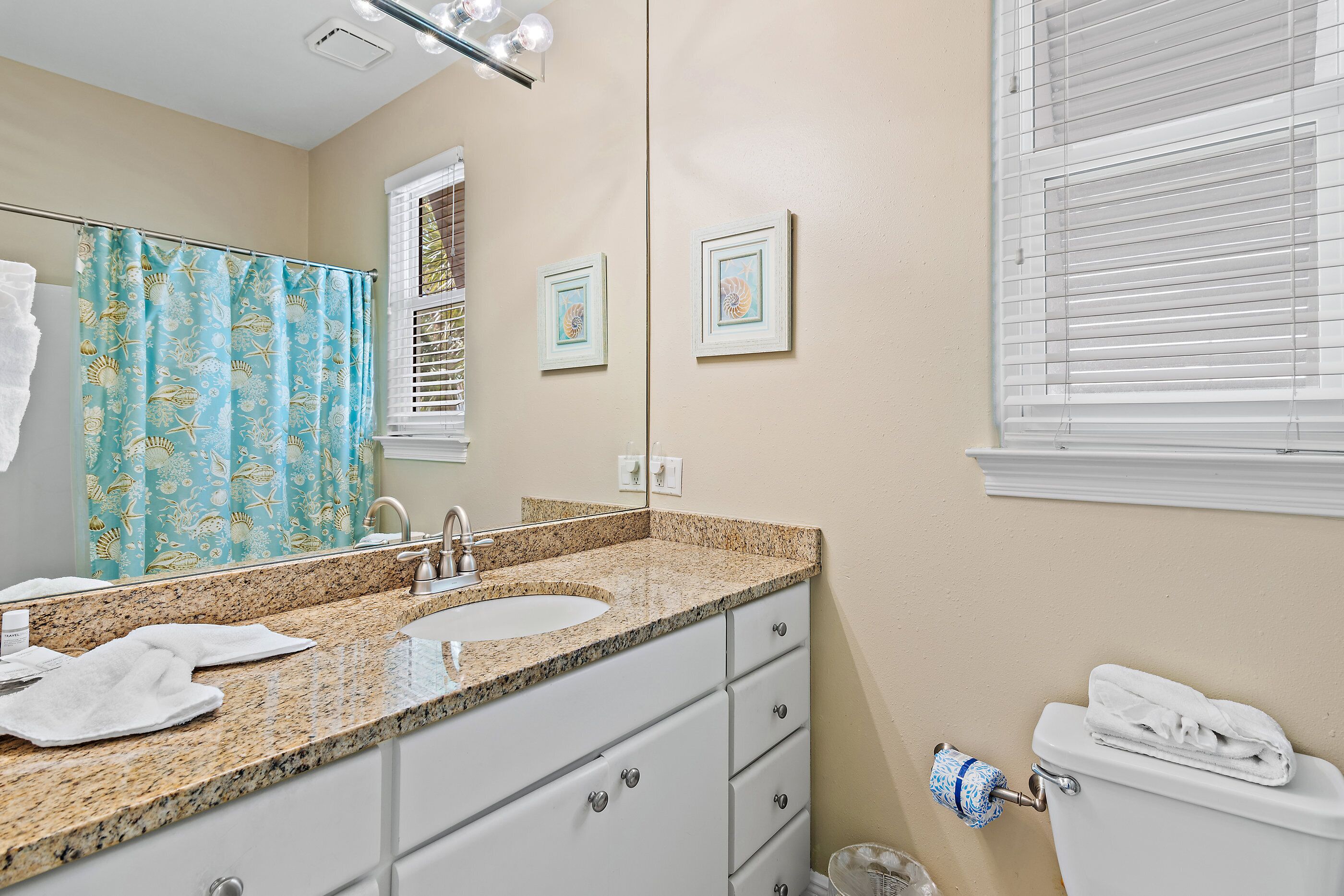 A bathroom with a granite countertop sink, a mirror, a toilet with a towel on top, and a shower curtain with a sea theme.