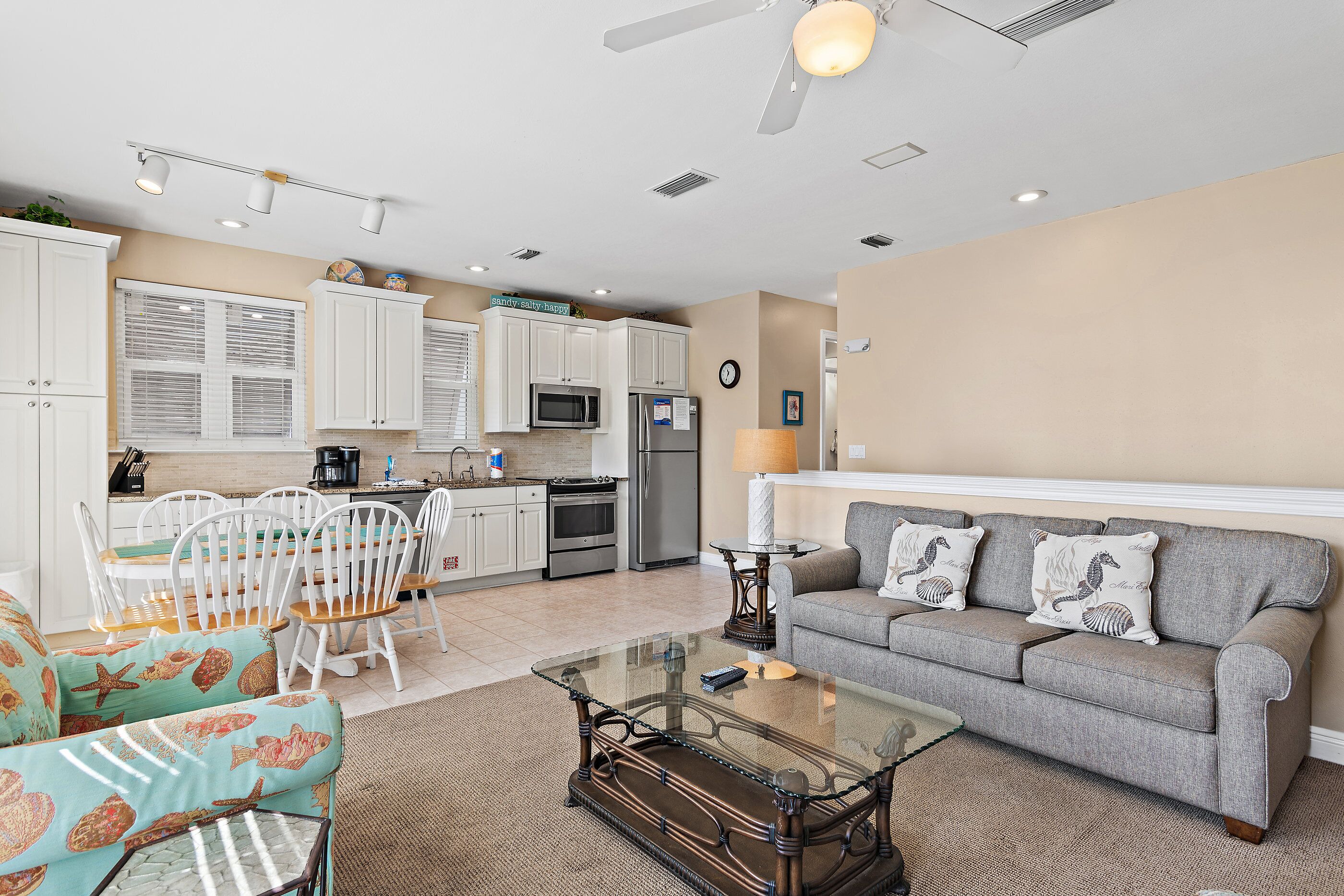 A modern kitchen with white cabinets, a dining area with white chairs, a grey sofa, and a glass coffee table.
