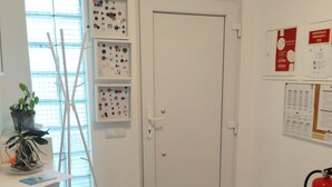 Una habitació amb una porta blanca, una finestra amb persianes, una estantenca penjada a la paret amb diversos objectes, un extintor vermell i una catifa marró a terra.